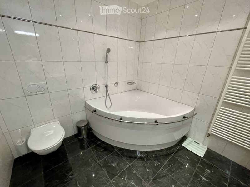 Modern bathroom with a white bathtub, toilet, and a chrome faucet. The walls are tiled in white with a dark marble floor.