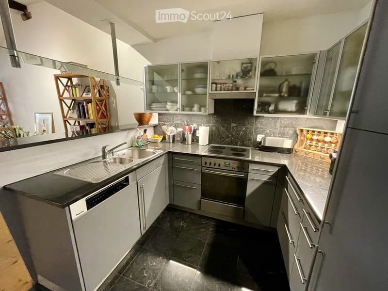 Modern kitchen with stainless steel appliances, dark countertops, and grey cabinets. Features include a sink, oven, dishwasher, and a small shelf with decorative items.