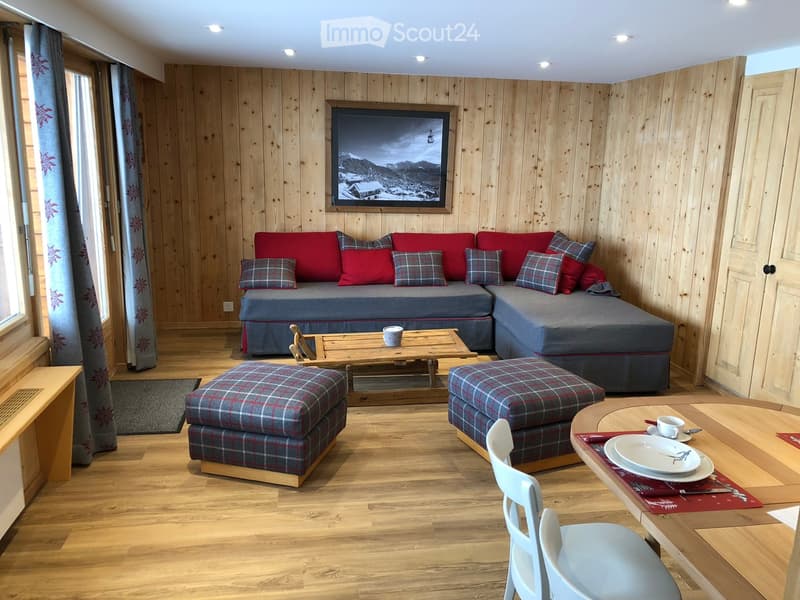 Cozy living room with wooden walls and floor, red sofa with plaid cushions, coffee table, ottomans, dining table with chairs, and a scenic mountain view picture on the wall.