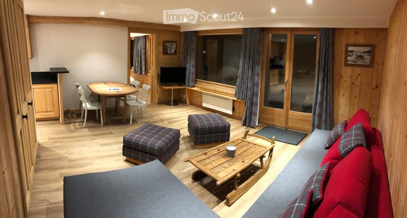 Cozy living room with wooden floors, a red sofa, coffee table, ottomans, dining area with a table and chairs, and a television. Large windows and wooden walls.