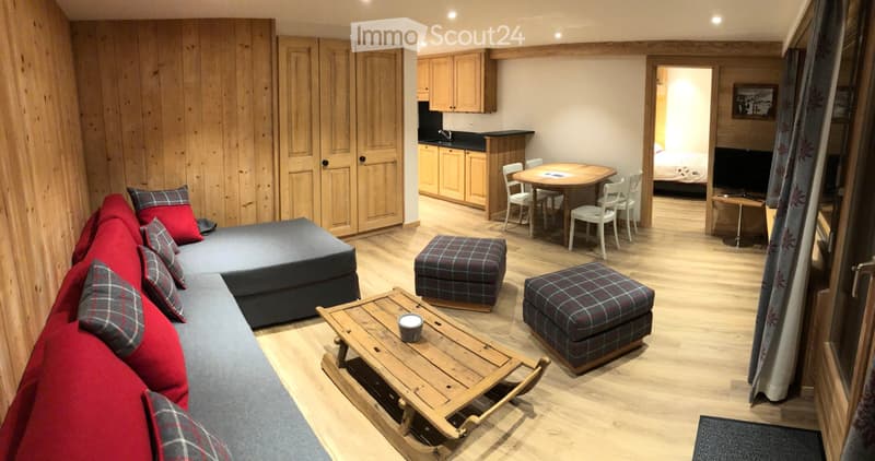A cozy, open-plan living and dining area with wooden floors, a grey sectional sofa with red and plaid pillows, a wooden coffee table, ottomans, a dining table with chairs, and a small kitchen area with cabinets and a sink.