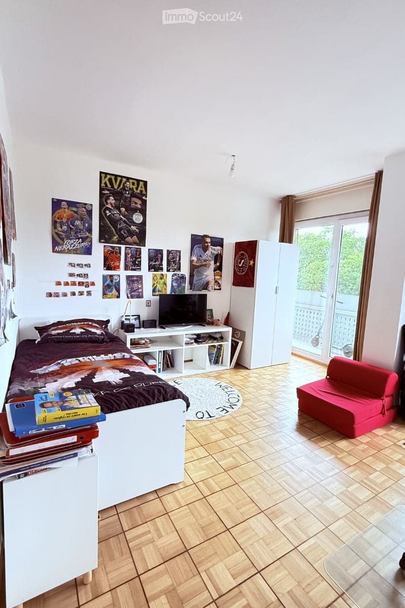 Cozy bedroom with a bed, sofa, TV, bookshelf, and a balcony access. Decorated with sports-themed posters and a round rug.