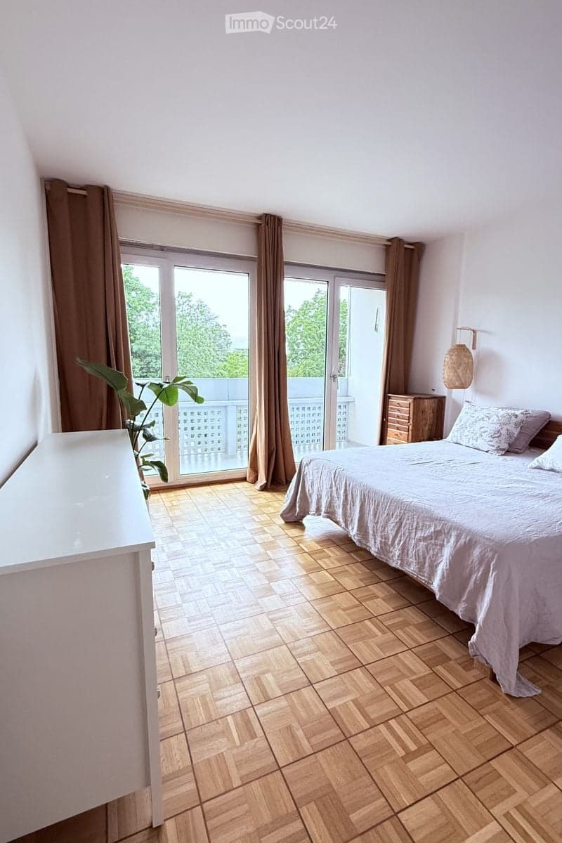 Bright bedroom with wooden herringbone flooring, large sliding glass doors leading to a balcony, a neatly made bed with pillows and a duvet, a nightstand, and a potted plant near the window.