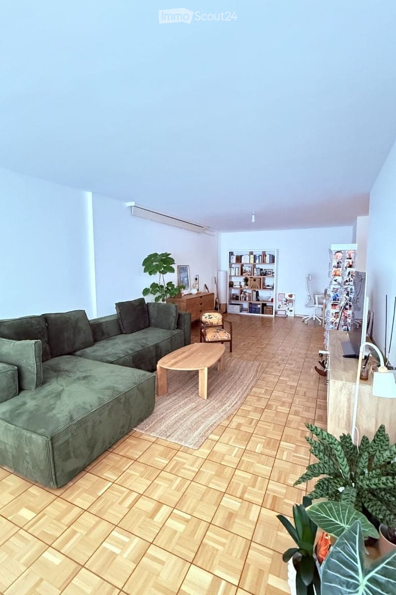Modern living room with a green sectional sofa, wooden coffee table, armchair, bookshelf, and plants. The room features wooden flooring and large windows.