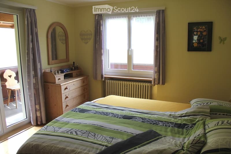 Bright bedroom with a double bed, dresser, mirror, chair, and a window with curtains. The room features a balcony access and a radiator under the window.