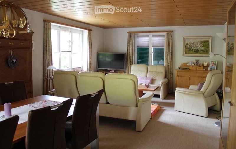 Bright living room with light-colored furniture, including a sofa, chairs, a coffee table, and a television. The room features large windows with curtains, a wooden ceiling, and a dining area with a table and chairs.