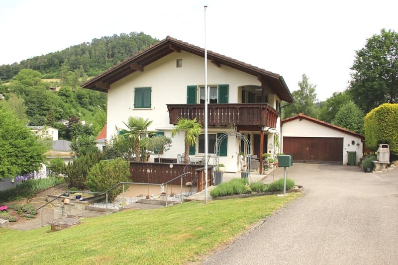 Two-story house with a balcony, garage, and well-maintained garden. The house features green shutters and a wooden balcony railing.