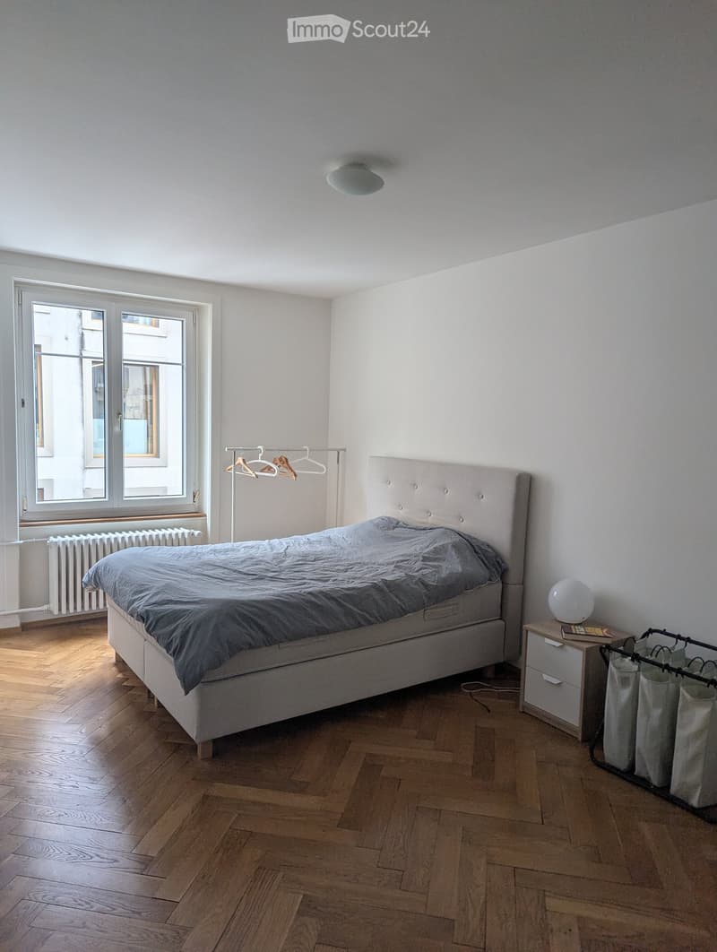 Minimalist bedroom with a single bed, nightstand, lamp, radiator, and a hanger rack near the window.