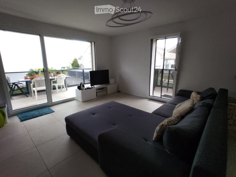 Modern living room with large sliding glass doors leading to a balcony, a dark grey sectional sofa, a small white TV stand, and a round table with chairs on the balcony.
