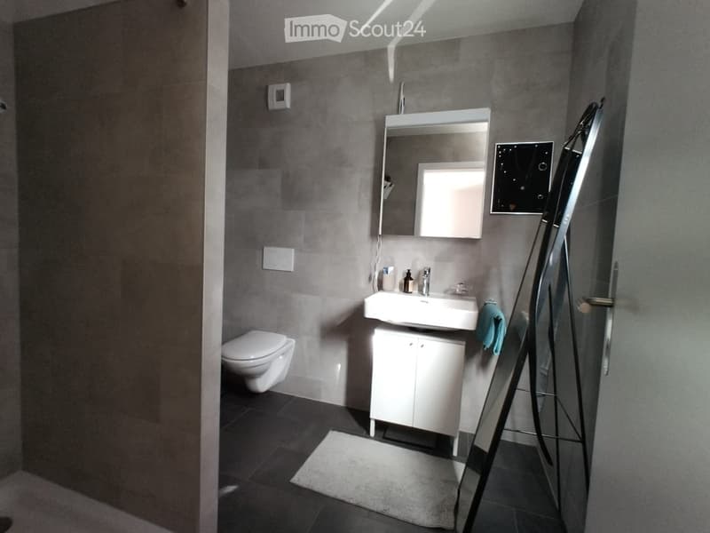 Modern bathroom with grey tiled walls, white sink, mirror, toilet, and a small cabinet. A towel hangs on the side of the sink.