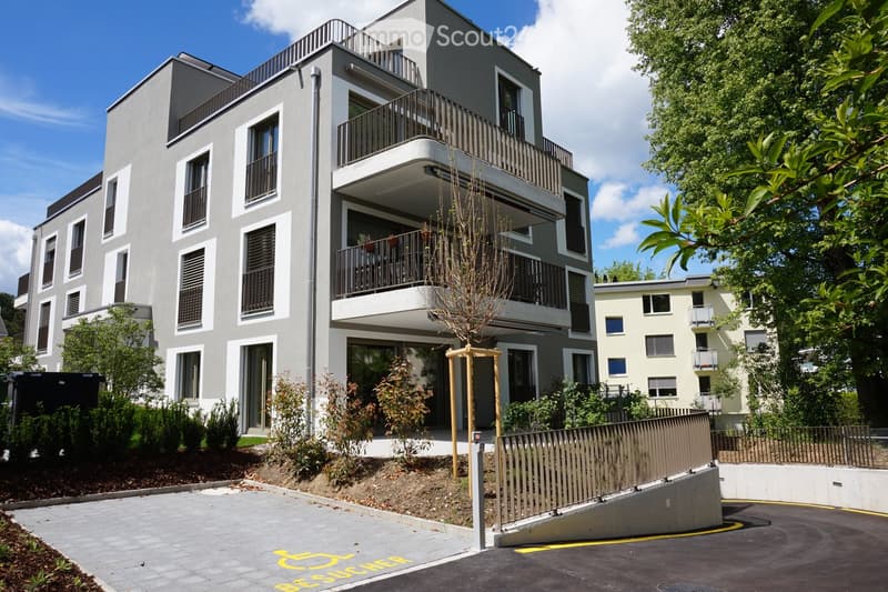 Modern multi-story residential building with balconies, large windows, and contemporary design. The building features a raised ground floor and is surrounded by greenery and a paved parking area.