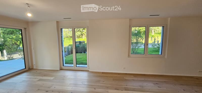 Empty room with light wooden flooring, white walls, large windows, and sliding glass doors leading to a green outdoor area.