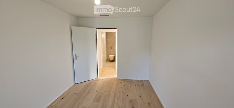 Empty hallway with light wooden flooring and white walls, leading to a small bathroom with a toilet and tiled walls.