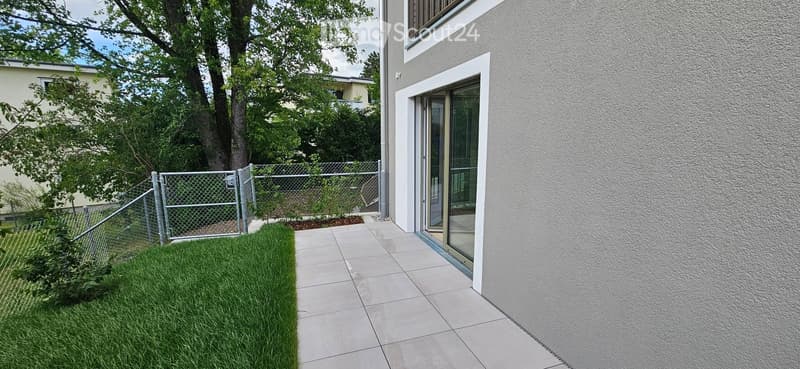 A small, modern garden area with a neatly trimmed lawn, a metal fence, and a tiled pathway leading to a house entrance with large glass doors.