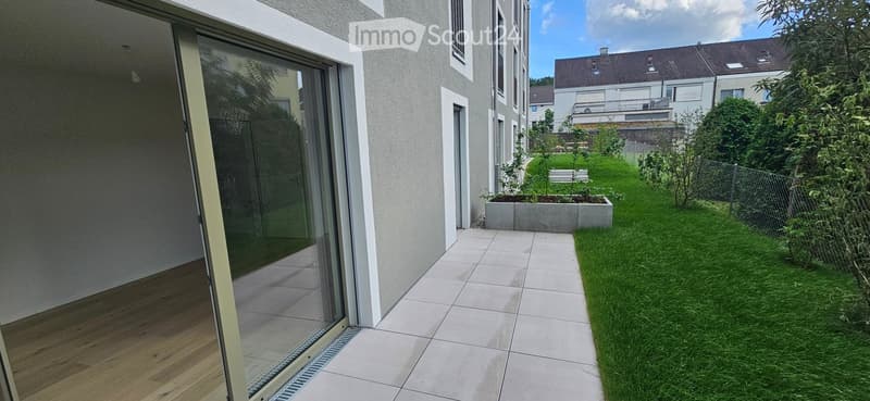 Modern outdoor patio area with tiled flooring, glass sliding door leading to interior, small garden with grass, plants, and a raised planter box.