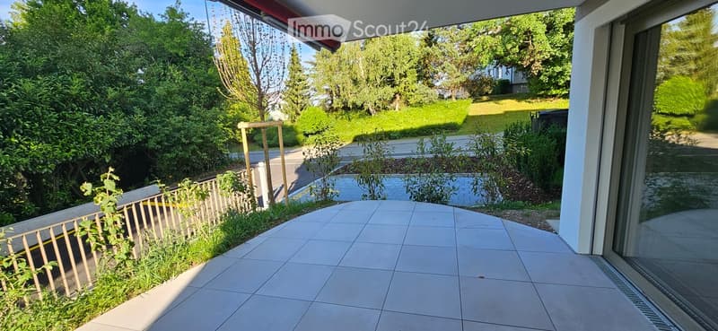 Balcony area with tiled flooring, wooden railing, and surrounding greenery, including bushes and trees.