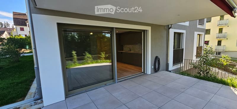 Balcony with tiled floor, sliding glass doors leading to a kitchen area, greenery in the background, and neighboring buildings visible.
