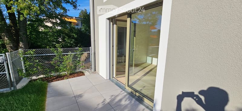 Modern balcony with large glass sliding doors, tiled floor, and a small fenced garden area with grass and plants.