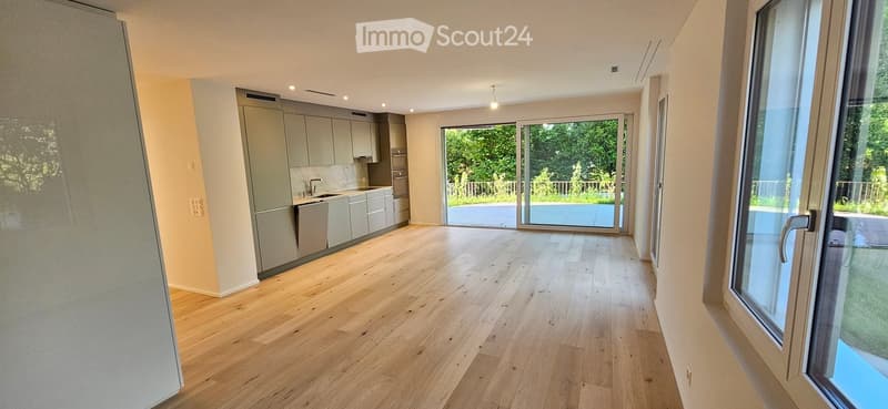 Open-plan kitchen and living area with light wooden flooring, modern grey cabinets, and large glass sliding doors leading to a garden view.