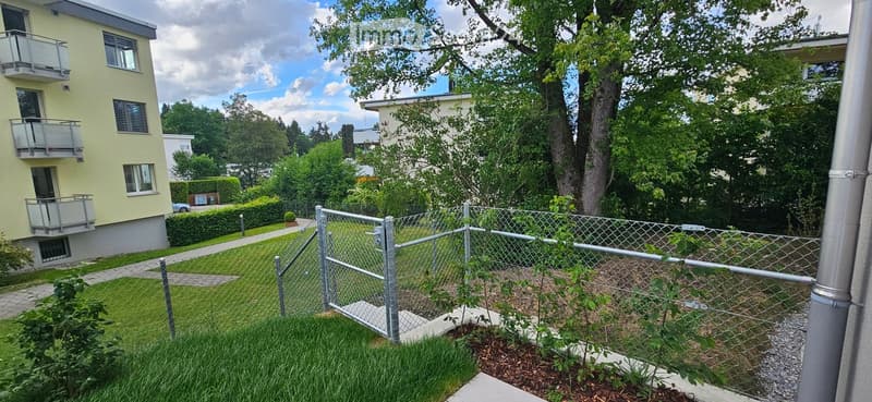 A fenced garden area with a well-maintained lawn, small plants, and a pathway leading to a multi-story residential building with balconies.
