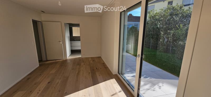 Empty room with wooden flooring, large sliding glass door leading to a patio, and visible adjacent bathroom with white cabinets and sink.