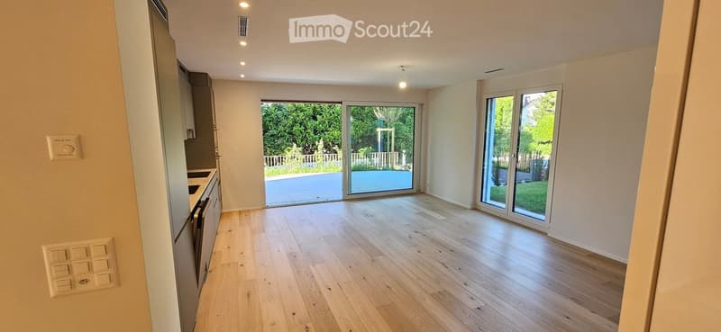 Bright, empty room with light wooden flooring, large sliding glass doors leading to a garden, and modern kitchen cabinets on one side.