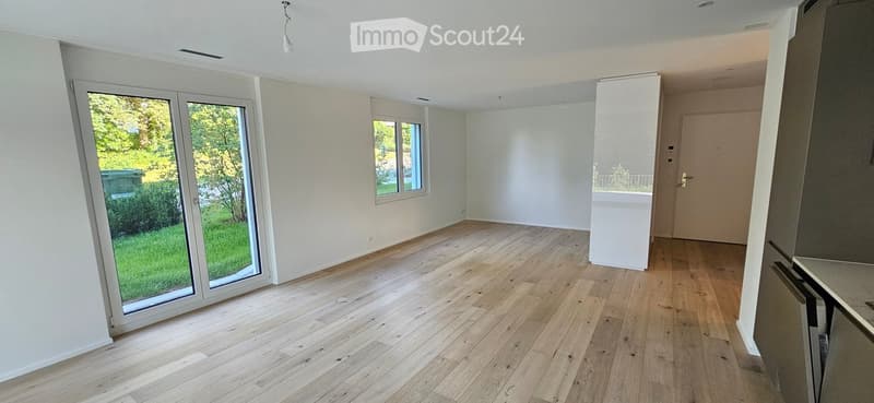 Empty room with light wooden flooring, white walls, large sliding glass door leading to a green outdoor area, and a small window on the opposite wall.