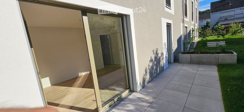 Modern balcony with large glass sliding doors, tiled floor, and adjacent grassy area with low concrete planters.