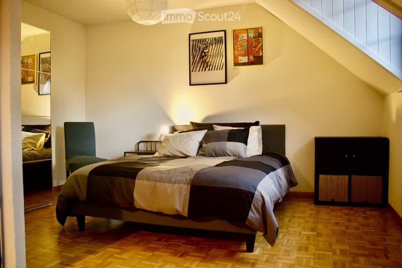 Cozy bedroom with a large bed, nightstand, chair, and cabinet. The room features wooden flooring, white walls, and modern decor including artwork and a mirror.