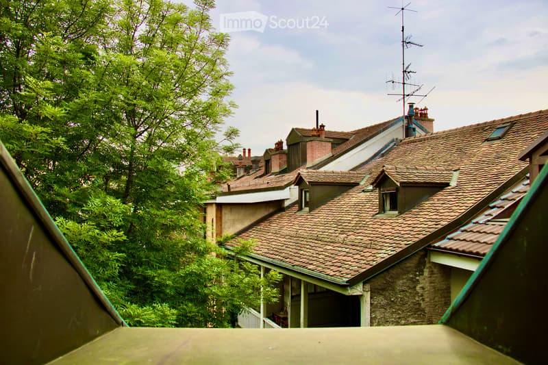 A view of several residential buildings with red-tiled roofs, chimneys, and an antenna on one roof, surrounded by lush green trees.