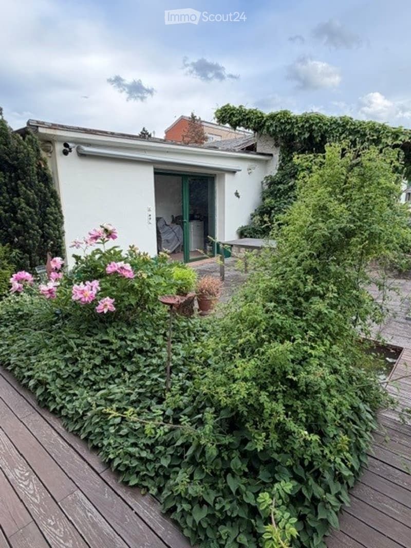 A lush garden area with a wooden deck, various plants, flowers, and a small seating area near the entrance of a house.