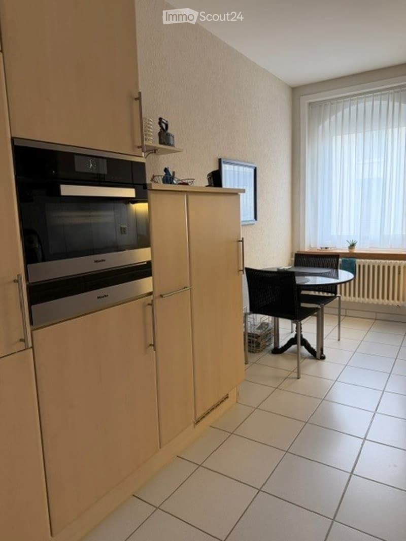 Modern kitchen with light wooden cabinets, built-in oven, tiled floor, and a small dining area with a table and chairs.