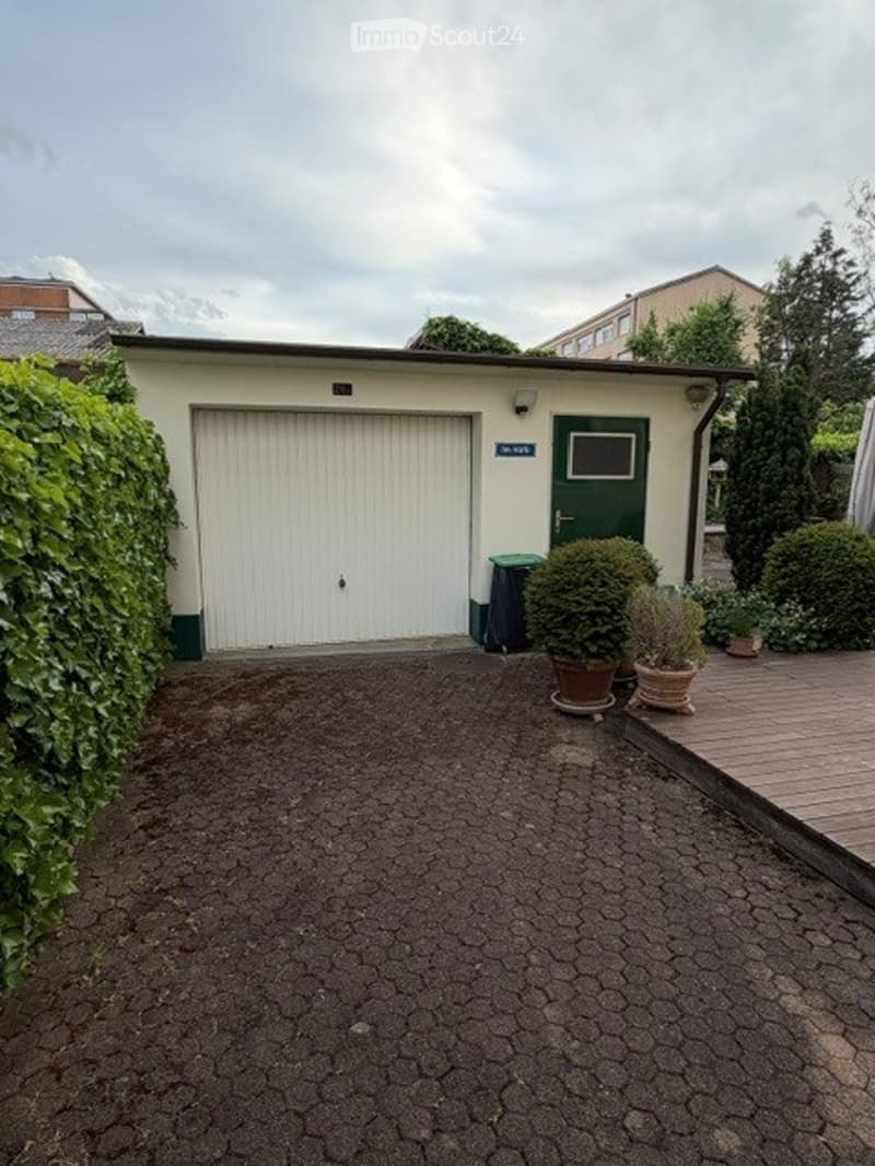 Small garage with a white door, surrounded by greenery and potted plants, with a paved driveway leading to it.