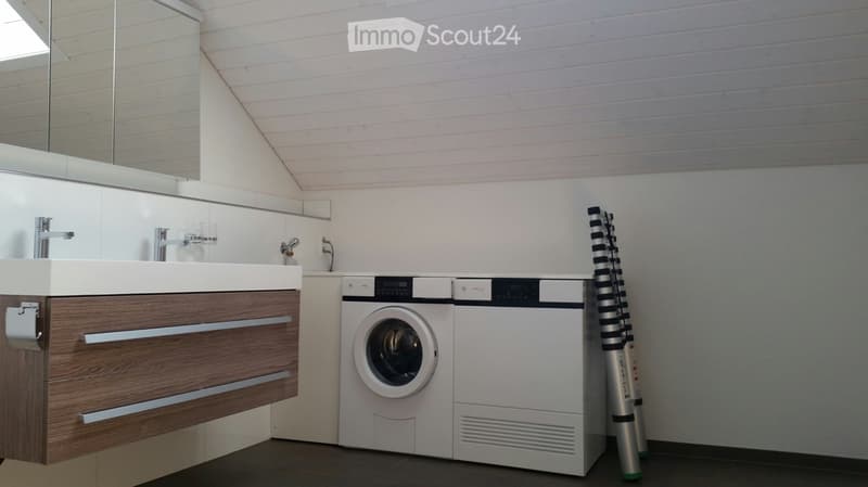 Modern bathroom with a sleek wooden vanity, integrated sink, and a compact washer/dryer unit. A large mirror and a tall, striped stool are also visible.