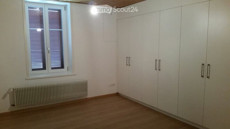 Empty room with light-colored walls, wooden flooring, built-in white wardrobes, a window with blinds, and a radiator below the window.