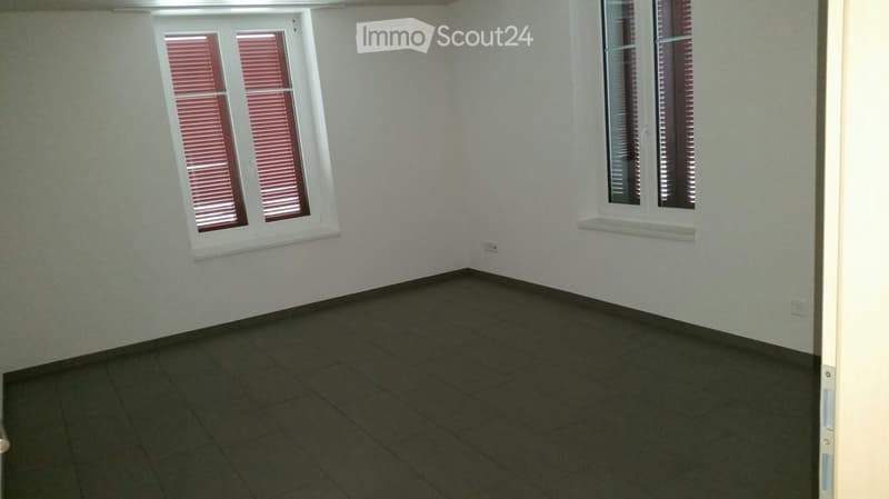 Empty room with light-colored walls, dark tiled floor, and two windows with red blinds.