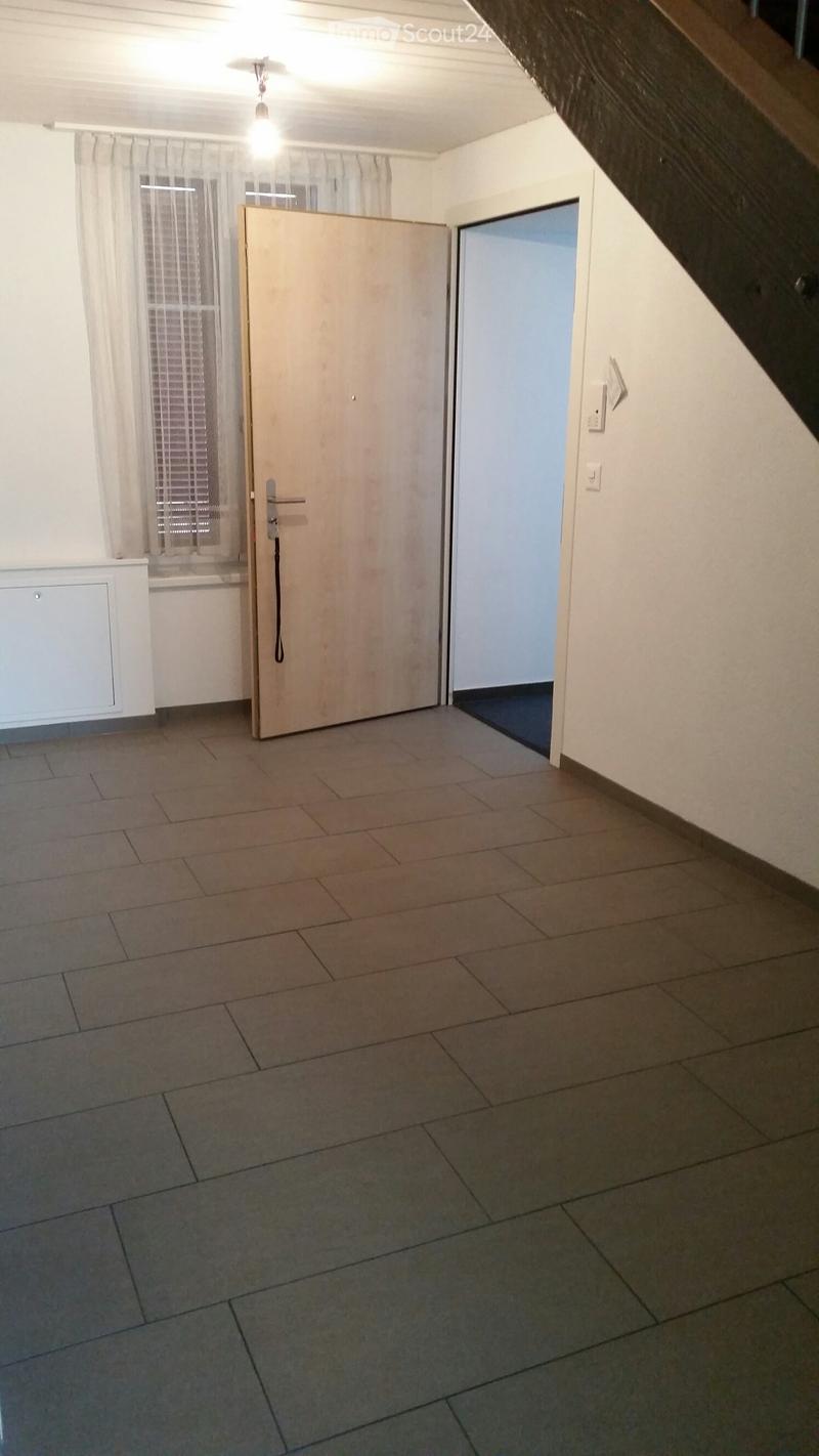 Empty room with tiled floor, white walls, and a closed wooden door leading to another space. A window with blinds is visible on the left side.