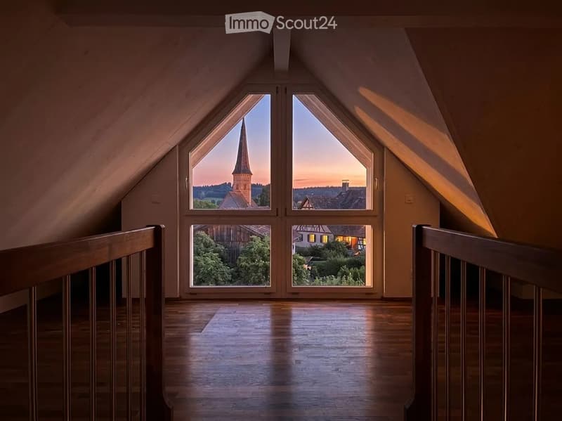 Bright, empty attic space with wooden floors, large windows offering a scenic view of a town and countryside.