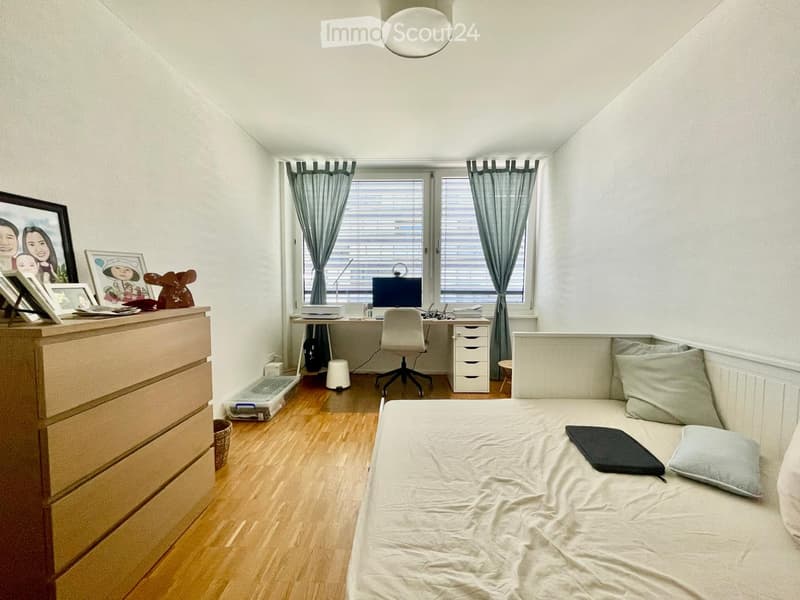 A cozy bedroom with a bed, desk, and chair. The room features a wooden floor, white walls, and large windows with curtains. A dresser with picture frames and a small stool are also visible.