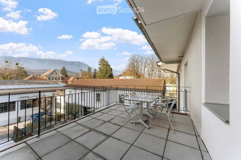 Balcony with tiled floor, metal railing, and a small table with chairs, overlooking a scenic view of mountains and houses.