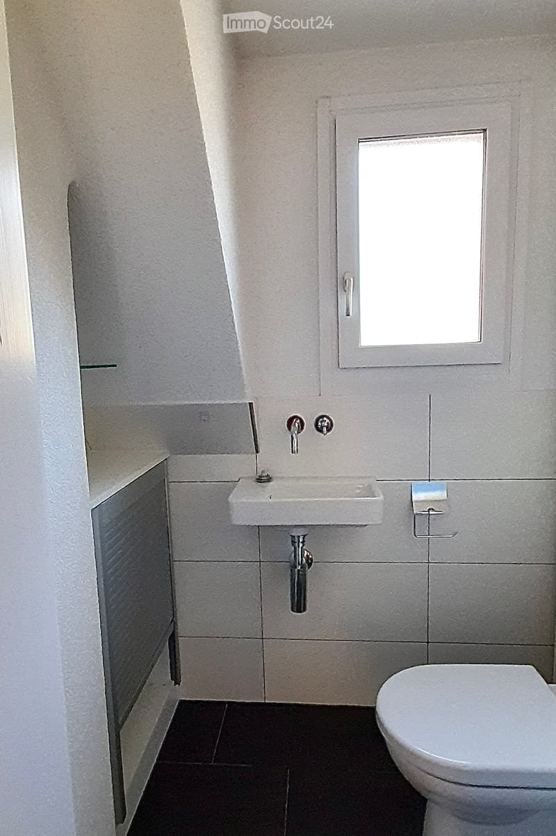 Modern bathroom with white tiled walls, dark tiled floor, a single sink with a chrome faucet, and a toilet. A small window provides natural light.