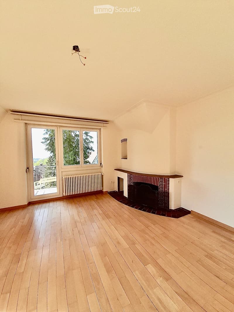 Empty room with wooden flooring, white walls, a fireplace with a brick surround, a radiator, and large windows with a view of greenery outside.