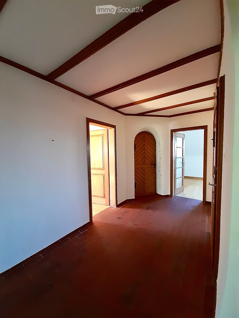 Empty hallway with wooden doors and red tiled floor, featuring white walls and wooden ceiling beams.