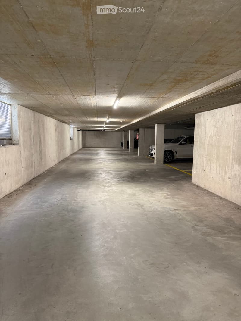 Underground parking area with concrete walls and ceiling, a few parked cars, and fluorescent lighting.