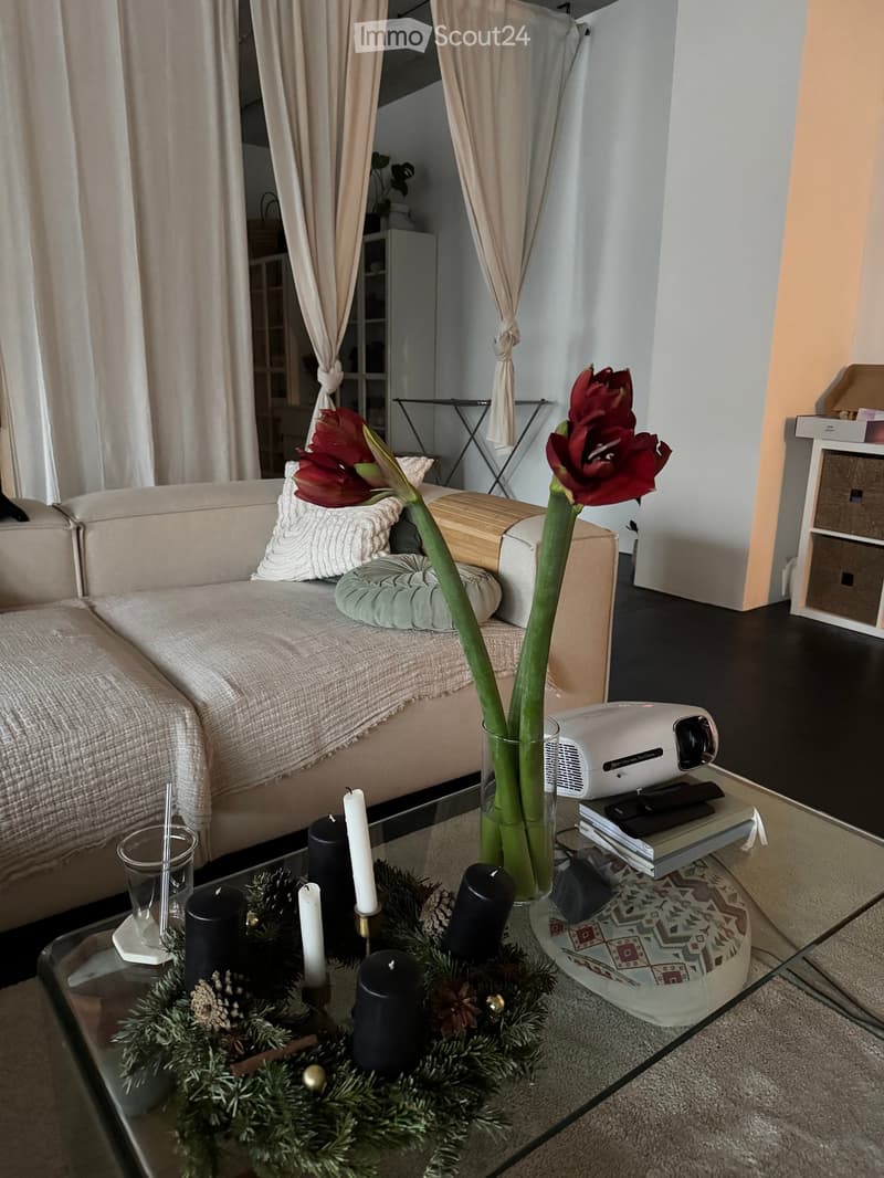 A cozy living room with a sofa, coffee table, projector, and decorative candles. A vase with red flowers sits on the coffee table, and a shelf is visible in the background.