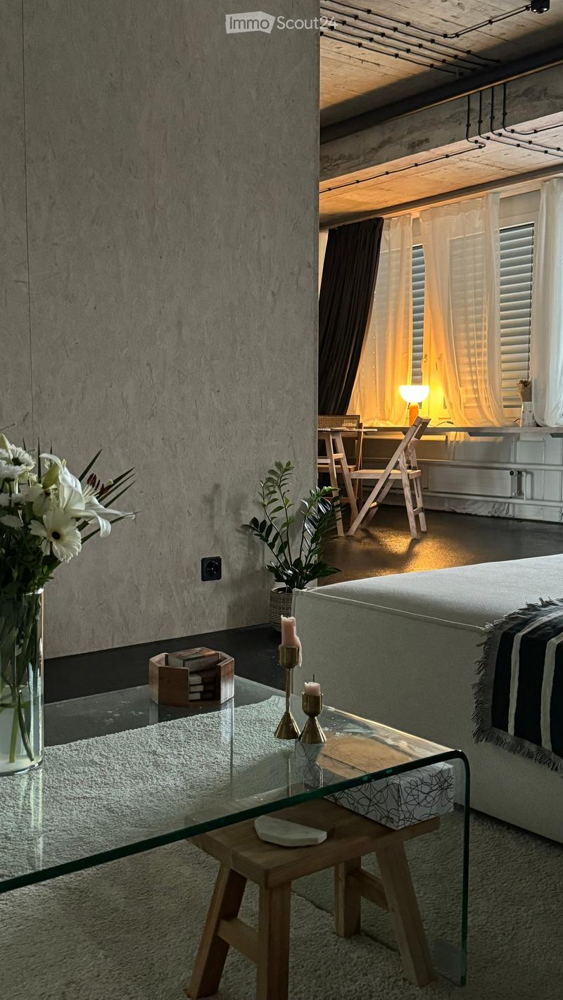 Modern bedroom with a grey bed, glass coffee table, wooden stool, vase with white flowers, candles, and a small workspace area with a chair and table.