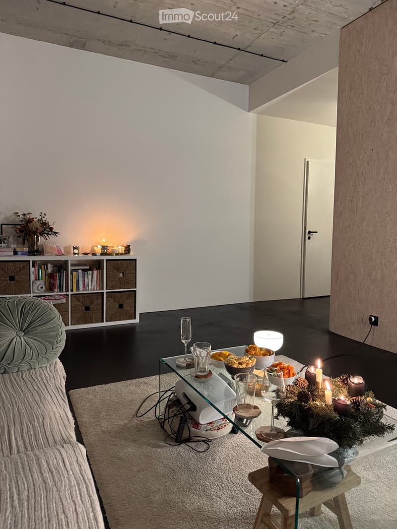 Modern living room with a dark wooden floor, white walls, and a high exposed wooden ceiling. Features a grey sofa, glass coffee table with decorative items, bookshelf, and a small wooden stool.
