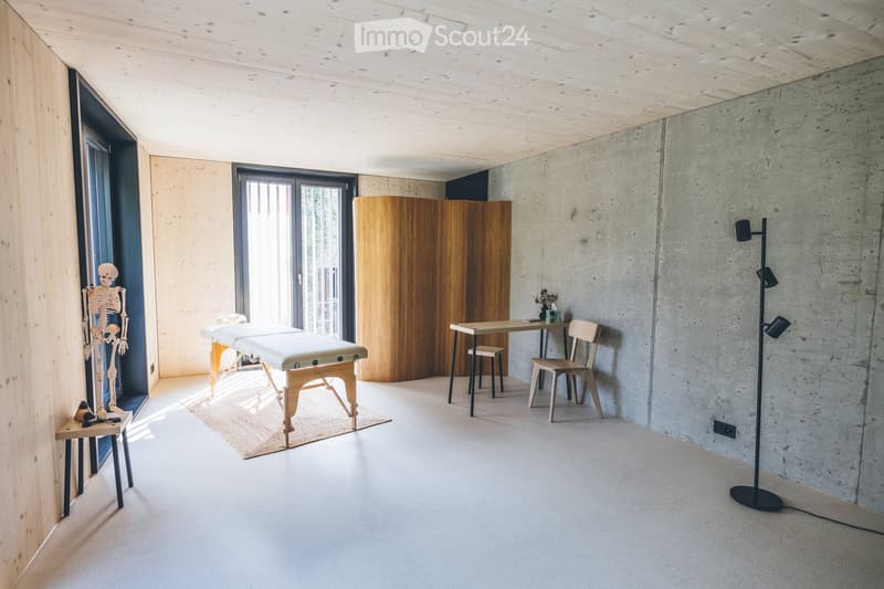 Modern, minimalist room with a massage table, wooden chair, small table, stool, skeleton model, floor lamp, and vase with flowers.