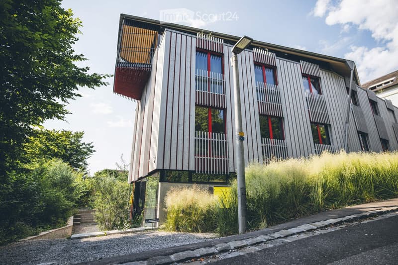 Modern two-story building with white and red exterior, large windows, balconies, and surrounding greenery.