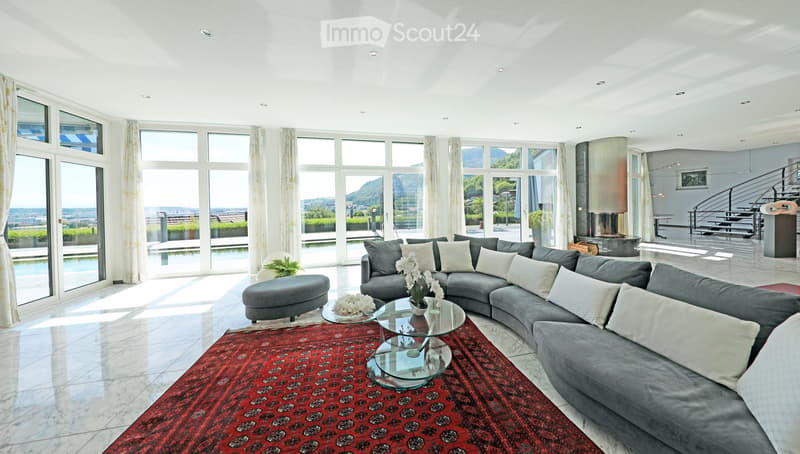 Modern living room with large windows offering a scenic view, featuring a grey sectional sofa, glass coffee tables, an ottoman, and a red patterned rug.